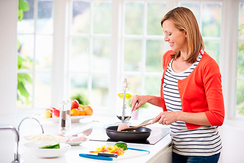 Smiling woman watches over a stove in a brightly lit kitchen